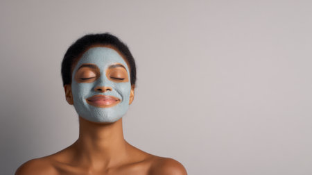 A serene young woman with closed eyes smiles as she relaxes with a blue clay face mask applied to her skin.の素材