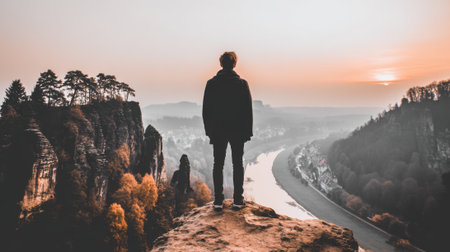 Rear view of a person on a rocky summit, gazing at a breathtaking sunrise over a winding river and misty mountains. A scene of adventure, freedom, and solitude.の素材