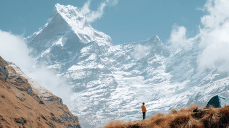 A lone traveler stands on a grassy precipice, dwarfed by the immense scale of a towering, snow-covered mountain under a cloudy blue sky, evoking a sense of adventure.の素材