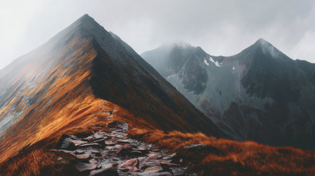Scenic view of a majestic mountain range on an overcast day. A rocky path winds along a colorful ridge, with foggy peaks creating a dramatic and atmospheric scene.の素材