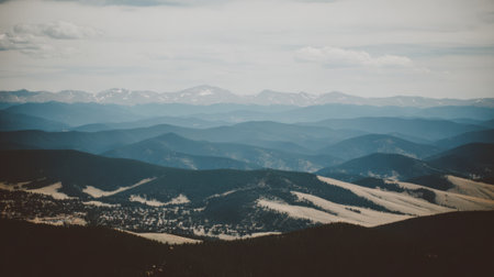 A breathtaking panoramic view of a mountain range with layers of hills fading into the distance under a cloudy sky. A small settlement is nestled in the valley below.の素材