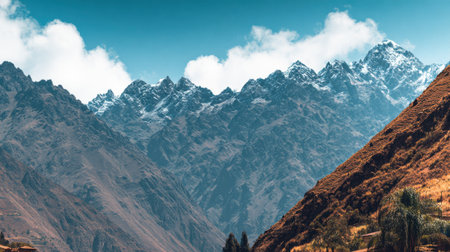 A stunning panoramic view of a rugged mountain range. The peaks are covered in snow against a bright blue sky with white clouds, with a rocky slope in the foreground.の素材