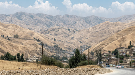 A panoramic landscape of dry, rugged mountains with a small town built on a slope. A paved road with cars and utility poles winds through the barren terrain under a partly cloudy sky.の素材