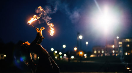 A skilled fire artist in silhouette performs a dangerous and beautiful dance with a flaming staff at night, with bright city lights creating a bokeh background.の素材