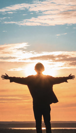 A backlit silhouette of a person standing with arms spread wide, facing a vibrant sunset. The scene evokes feelings of freedom, hope, and spiritual connection with nature.の素材