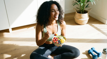 A beautiful, smiling Black woman in sportswear sits on the floor in a sunlit room, eating a healthy meal from a bowl after exercising at home. Wellness and self-care concept.の素材