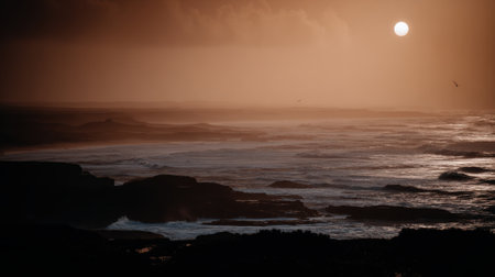 A tranquil and moody seascape featuring a hazy sun rising over a foggy ocean. Dark rocks are silhouetted in the foreground as gentle waves wash ashore in the soft light.の素材