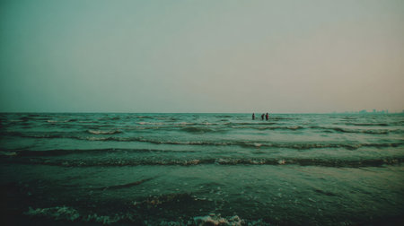 A wide shot of a vast, choppy ocean with a teal and green color palette. In the far distance, silhouettes of people can be seen in the water under a hazy, overcast sky.の素材