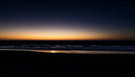 A tranquil beach at twilight with a vibrant orange and yellow sunset on the horizon. The calm ocean reflects the last light as stars begin to appear in the dark night sky.の素材