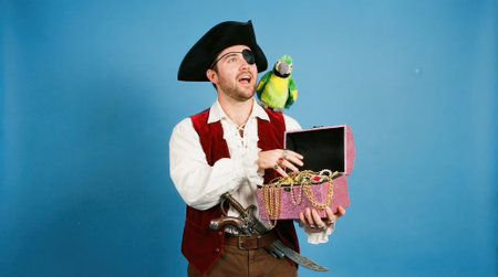 A man in a pirate costume with an eyepatch and hat smiles joyfully while holding an open treasure chest, with a colorful parrot perched on his shoulder against a blue background.の素材