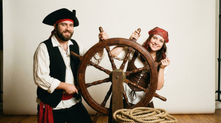 A smiling man and woman in fun pirate outfits with a hat and bandana stand behind a ships helm prop during a studio photoshoot against a white background.の素材