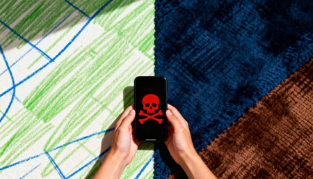 A person holds a smartphone displaying a red skull and crossbones symbol, indicating a digital threat or cybersecurity risk. The phone is centered against a background of green and blue patterned fabric, suggesting a casual, everyday setting where such threats can emerge.の素材