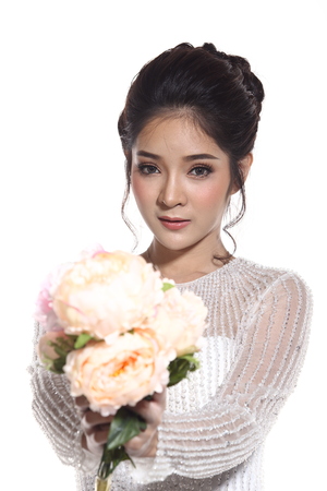 Portrait head shot with flower bouquet, Lovely Asian Beautiful Woman bride in white wedding gown dress with lace, black hair, studio lighting white background, isolated copy space, sweet romantic soft alone conceptの写真素材