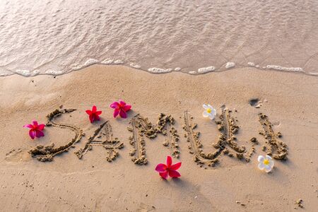 inscription on the sand of Samui, an island in Thailand, exotic frangipani flowers on the seashore, paradise plumeria flowers, background picture with flowers by the ocean, yellow sand in Asiaの写真素材