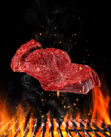 Flying raw steak above barbecue grill with fire flames. Concept of food preparation in low gravity mode. Separated on black backgroundの写真素材