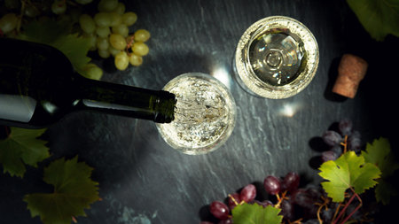 Top shot of pouring white wine into glass. Still life with grapes of wine served on dark stone...