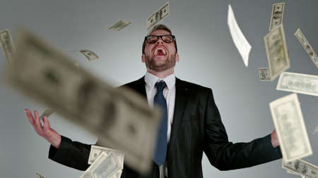 Close-up of screaming businessman with flying dollars banknotes isolated over gray backgroundの写真素材