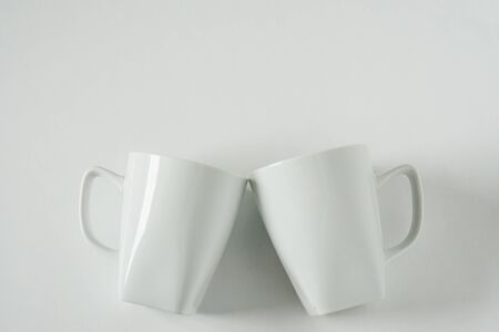 2 monochromatic white coffee mugs on white background clinking in cheers with blank empty room space for text, copy, or copy space. Modern top view concept of two cups with solid background backdrop.の写真素材