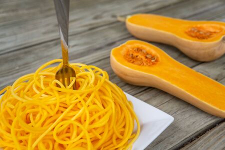 Butternut squash noodles with fork stuck in the middle of the pasta. Served on white plate on wooden background with cut in half vegetable in background. Low carb, keto, vegan, and gluten free trend.の写真素材