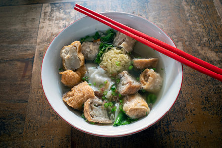 A bowl of meatball soup with fresh vegetables and red chopsticks on the old wooden table.