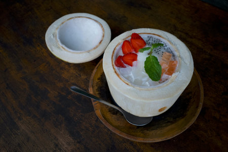 A mixed fruit salad, colorful dessert in half a fresh coconut, on a dark wooden table.
