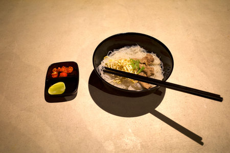 A bowl of ramen noodle with chopstick with sliced chili and sliced lime on a white table.
