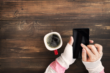 Above view of female hand holding smart phone with hot cup of coffee on wood table