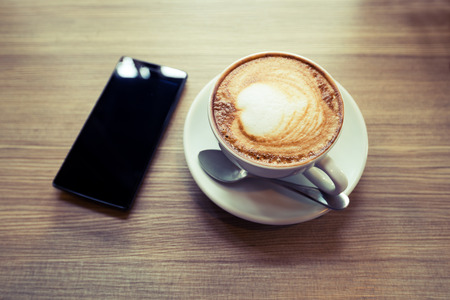 Above view of smart phone with hot cup of coffee on wood table. Photo in vintage color image style.の写真素材