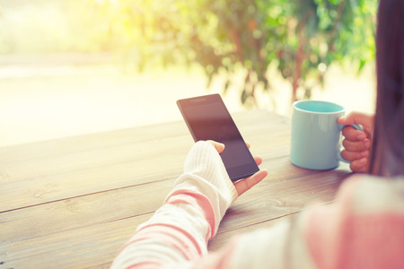 Rear view of female hand holding smart phone with hot cup of coffee on wood tableの写真素材