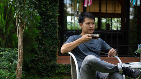 An adult Asian man wearing a gray shirt and jeans, sitting on a white shirt in the garden in front of the house, holding a cup of black coffee.の写真素材