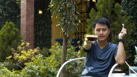 An adult Asian man wearing a gray shirt and jeans, sitting on a white shirt in the garden in front...