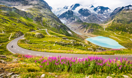Beautiful view of motorcyclist driving on winding mountain pass road in the Alps through gorgeous scenery with mountain peaks, glaciers, lakes and green pastures with blooming flowers in summerの写真素材
