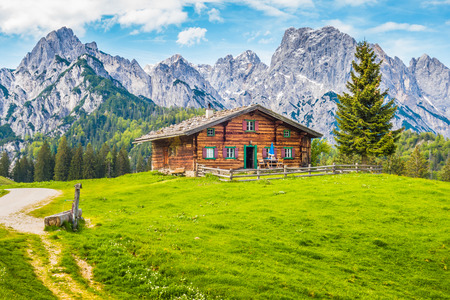 Panoramic view of scenic mountain landscape in the Alps with traditional old mountain chalet and fresh green meadows on a sunny day with blue sky and clouds in springの写真素材