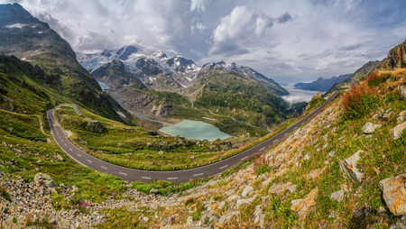 Beautiful view of motorcyclist driving on winding mountain pass road in the Alps through gorgeous scenery with mountain tops, ice glaciers, lakes and green pastures with blooming flowers on a sunny day in summerの写真素材