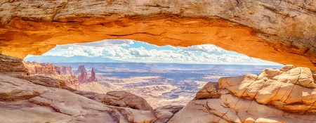 Panoramic view of famous Mesa Arch, iconic symbol of the American West, illuminated golden in beautiful morning light on a sunny day with blue sky and clouds, Canyonlands National Park, Utah, USAの写真素材