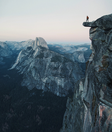 A fearless hiker is standing on an overhanging rock enjoying the view towards famous Half Dome at Glacier Point overlook in beautiful post sunset twilight, Yosemite National Park, California, USAの素材 [FY31073485132]