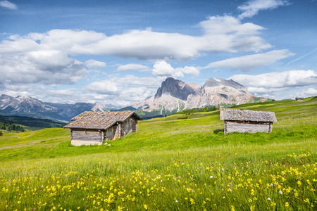 Beautiful view of idyllic alpine mountain scenery with traditional old mountain chalets and fresh green meadows on a sunny day with blue sky and clouds in springtime, Alpe di Siusi, South Tyrol, Italyの写真素材