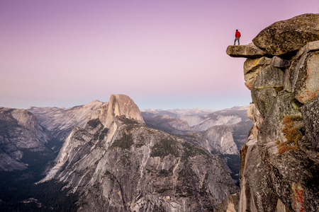 A fearless male hiker is standing on an overhanging rock at Glacier Point enjoying the breathtaking view towards famous Half Dome in beautiful post sunset twilight in summer, Yosemite National Park, Californiaの写真素材
