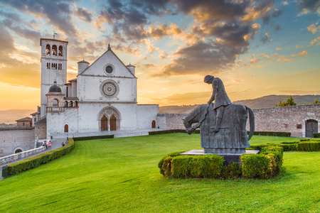 Classic view of famous Basilica of St. Francis of Assisi (Basilica Papale di San Francesco) with statue in beautiful golden evening light with dramatic clouds in the sky at sunset, Assisi, Umbria, Italyの写真素材