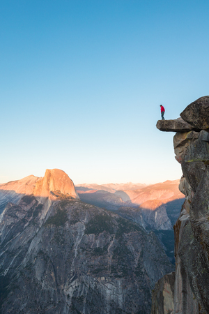 A fearless hiker is standing on an overhanging rock enjoying the view towards famous Half Dome at Glacier Point overlook in beautiful evening twilight, Yosemite National Park, California, USAの写真素材