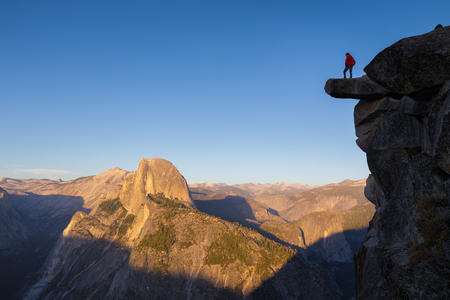 A fearless hiker is standing on an overhanging rock enjoying the view towards famous Half Dome at Glacier Point overlook in beautiful evening sunset twilight, Yosemite National Park, California, USAの写真素材