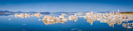 Panoramic view of fascinating tufa rock formations mirrored on calm water surface of famous Mono Lake on a beautiful sunny day with blue sky in summer, Mono County, California, USAの写真素材