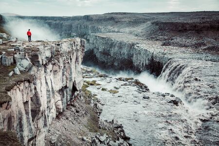 Panorama view of hiker in red jacket standing in front of gigantic Dettifoss waterfall on a moody day with dark clouds in summer, Skogar, Icelandの写真素材