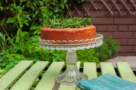 A perfect home baked sponge cake decorated with rosemary herb on crystal stand on green garden...