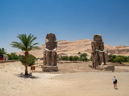 LUXOR, EGYPT : The Colossi of Memnon, two massive stone statues of Pharaoh Amenhotep III, stand on the west bank of the Nile, surrounded by desert landscape under a clear sky.の写真素材