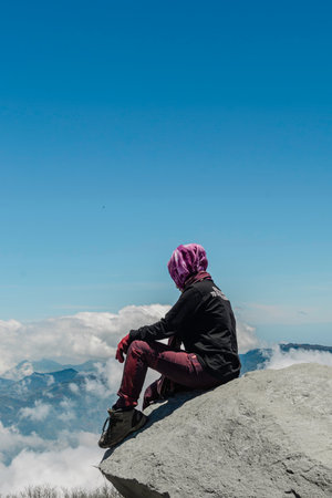 an Asian female hiker wearing a headscarf is sitting on a rock under a blue sky and on top of a mountainの写真素材