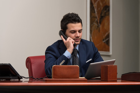 Young Businessman Working At His Computer While Talking On The Phoneの写真素材