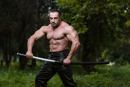 Action Hero Muscled Man Holding A Ancient Sword - Standing In Forest Wearing Leather Pantsの写真素材