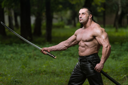 Action Hero Muscled Man Holding A Ancient Sword - Standing In Forest Wearing Leather Pantsの写真素材