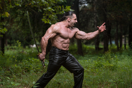Action Hero Muscled Man Holding A Ancient Sword - Standing In Forest Wearing Leather Pantsの写真素材
